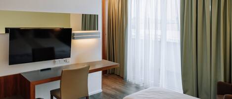 Una habitación de hotel con cama, escritorio con televisor, silla y ventana con cortinas.