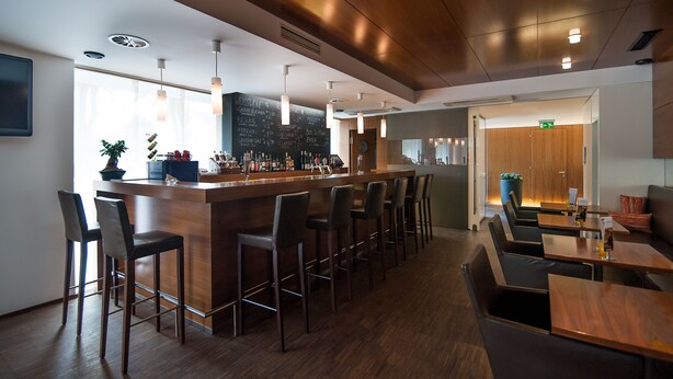 A modern bar area with wooden bar stools, a long counter, and pendant lighting. - HEINhotel Vienna Airport (Schwechat)