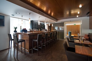 A modern bar area with wooden bar stools, a long counter, and pendant lighting. - HEINhotel Vienna Airport (Schwechat)