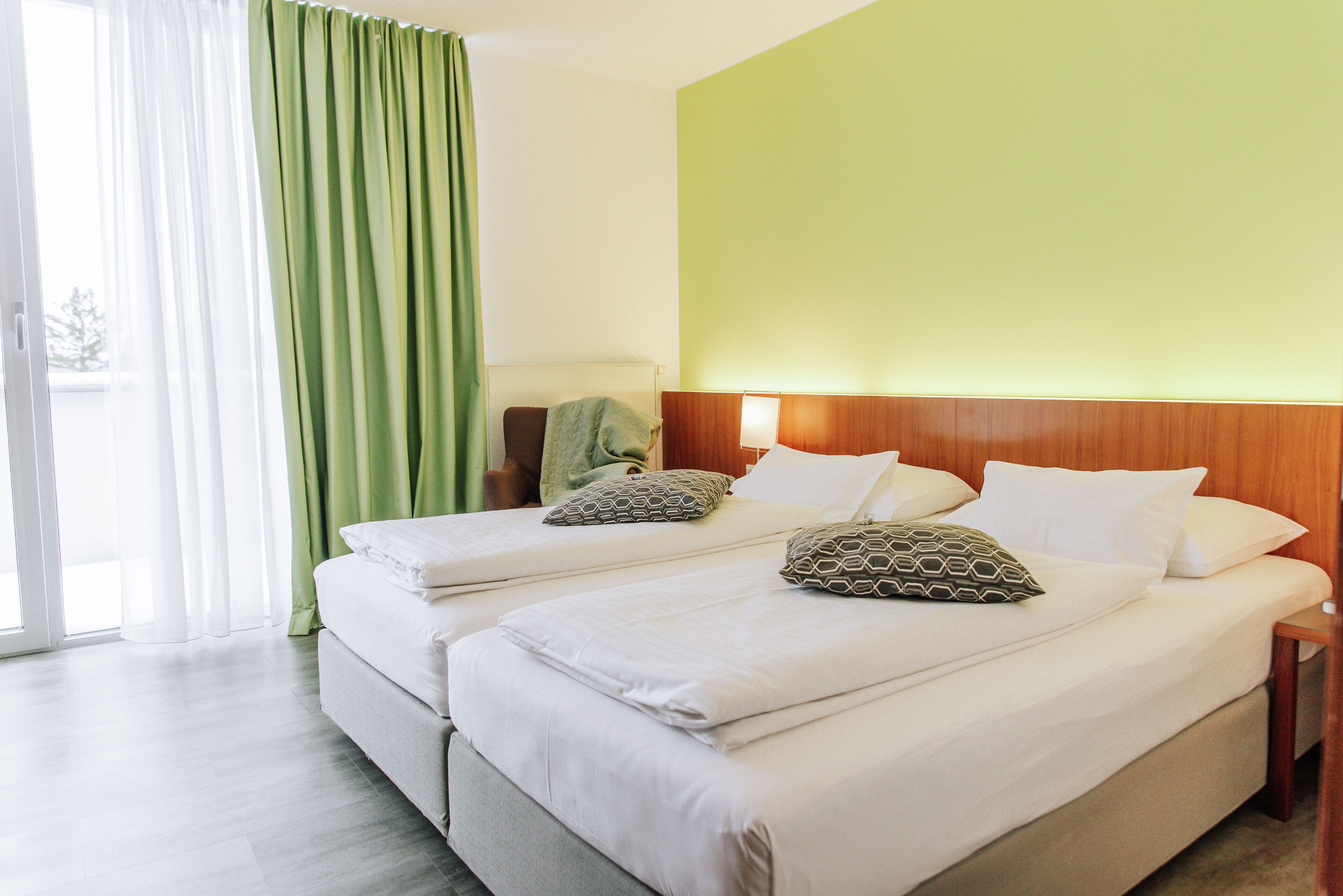 Une chambre d’hôtel avec deux lits, une tête de lit en bois, un rideau vert et une vue par la fenêtre.
