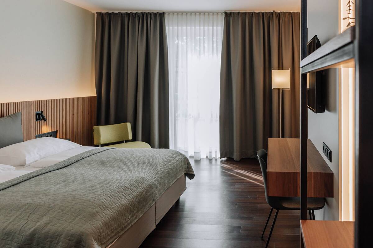 A hotel room with a large bed, a desk, a chair, a TV, and a window with curtains.