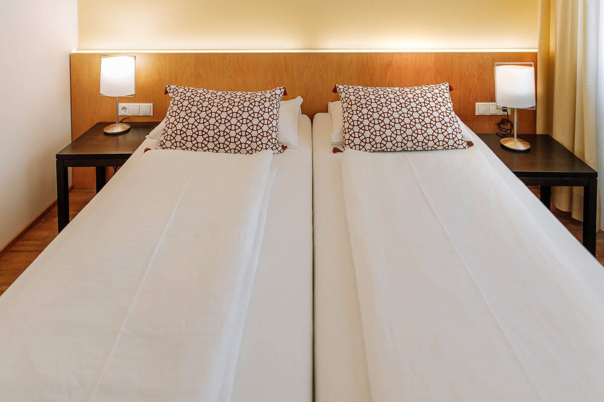 A double bed with white linens and patterned pillows, flanked by nightstands with lamps, in a hotel room.