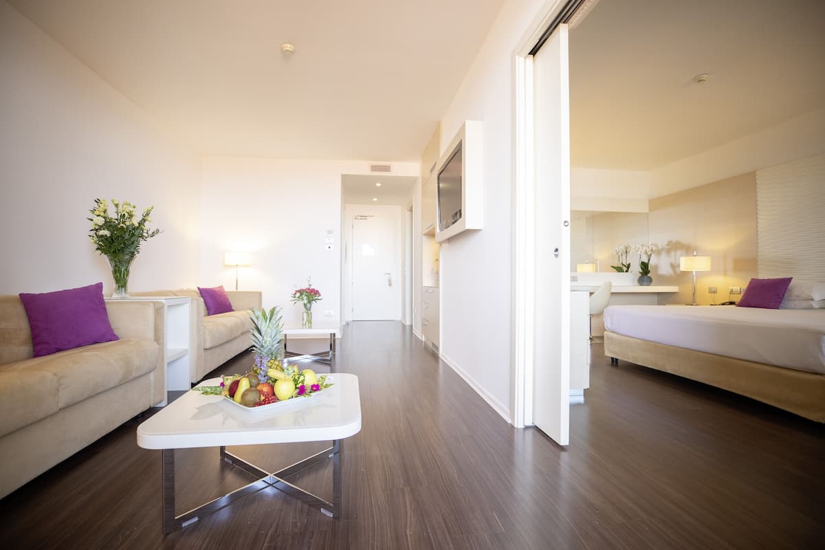 Una habitación de hotel moderna con sofá, una mesita de centro con fruta, cama y zona de escritorio.