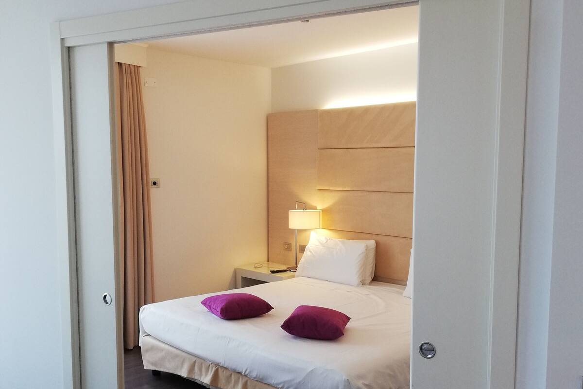 Una habitación de hotel moderna con una cama, una mesita de noche, una lámpara y una puerta que da a otra habitación.
