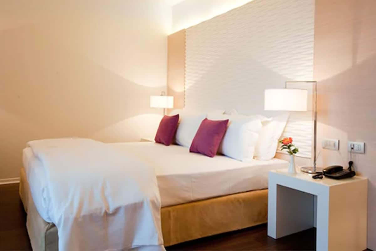 Una cama bien tendida con almohadas blancas y moradas, una lámpara de noche y un teléfono sobre una mesita de noche blanca.
