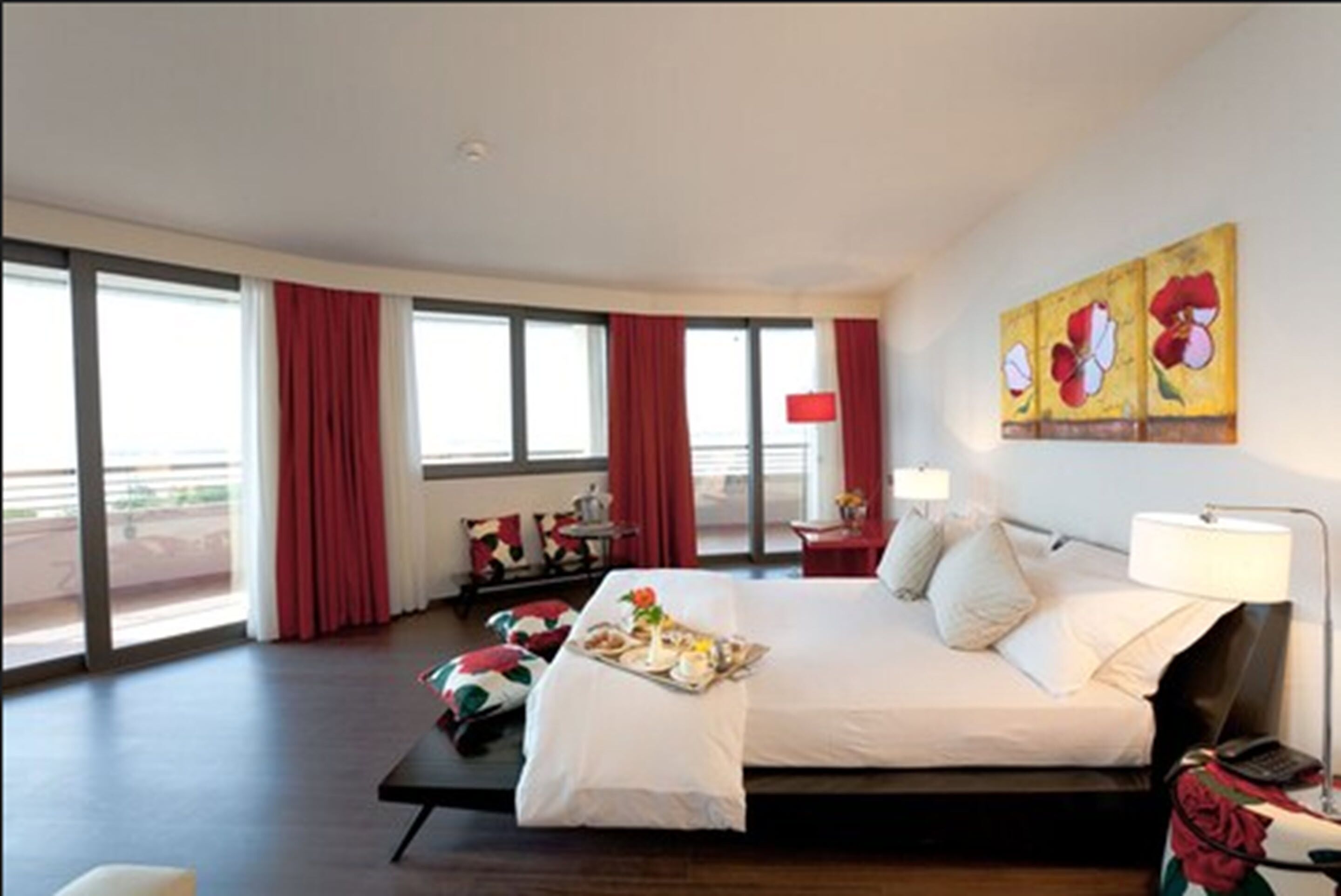 A modern hotel room with a large bed, a dining table with a tray of food, and a view of the outdoors through large windows.