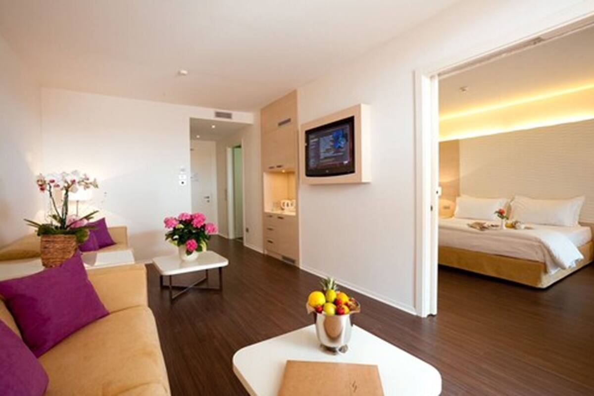 Una habitación de hotel moderna con una cama, una mesita con un frutero, un sofá con cojines morados y un televisor montado en la pared.
