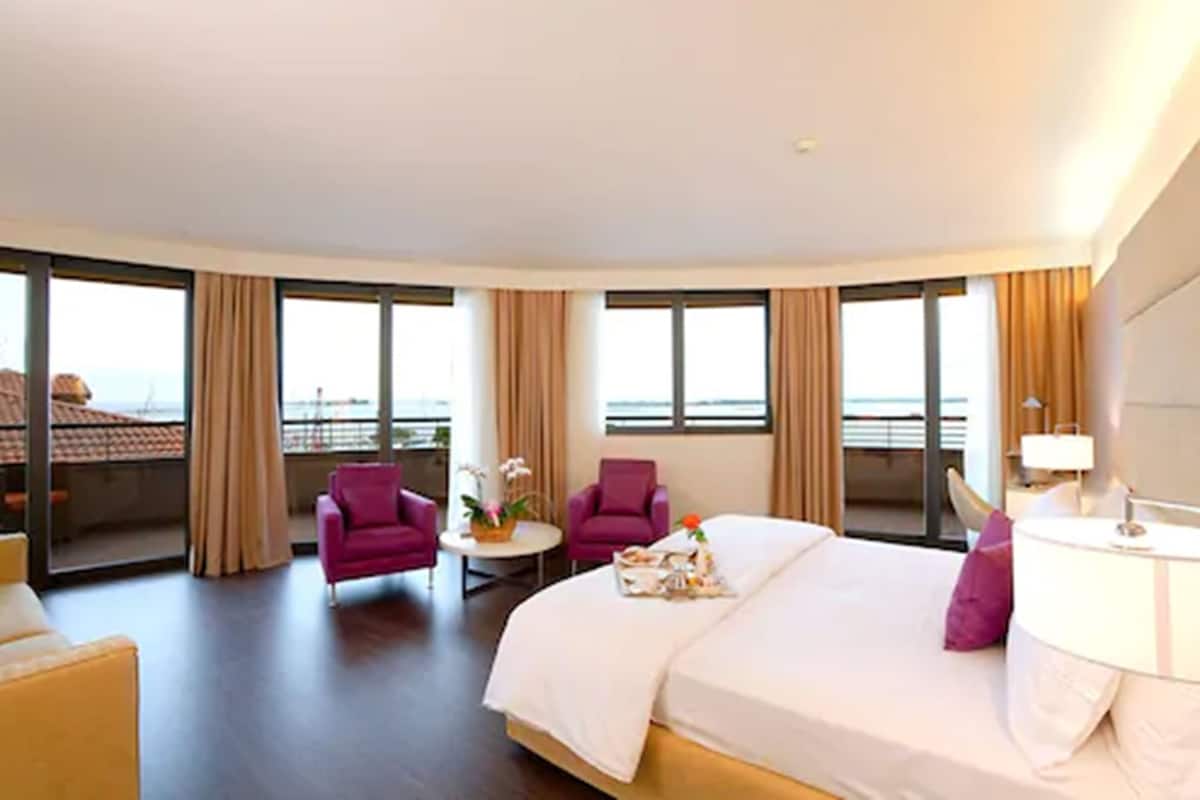 Habitación de hotel con una cama grande, dos sillones, una mesita con un florero y ventanas amplias que ofrecen una vista.