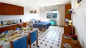 A compact hotel room with a kitchen area, a dining table with chairs, a bed with a striped bedspread, and a view of the city through the window. - Residence Edy (Imperia)