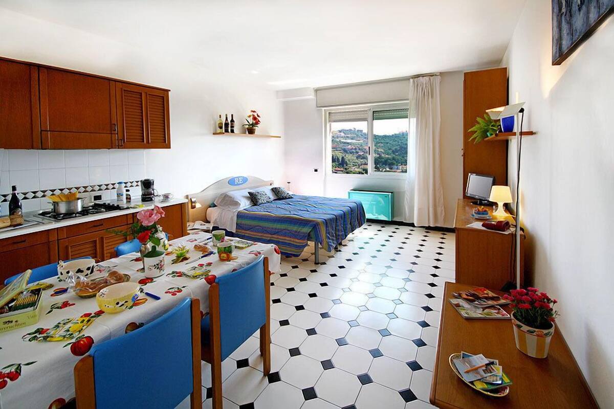 Habitación de hotel pequeña con cocina, una mesa de comedor con sillas, una cama con colcha a rayas y vistas a la ciudad desde la ventana.