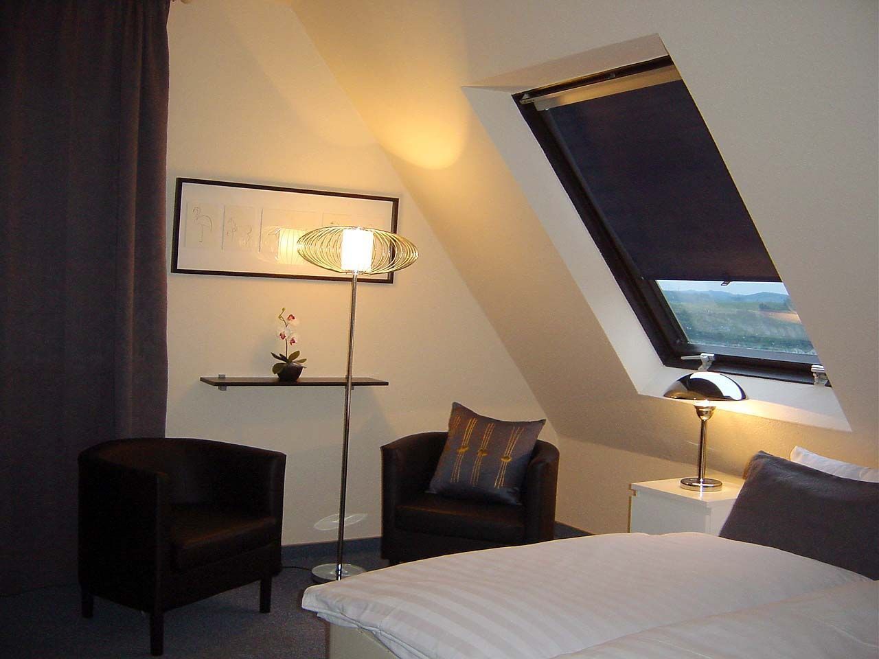 A hotel room with a bed, two chairs, a nightstand, a floor lamp, a shelf with a plant, and a window with a view.