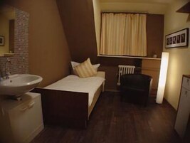 Habitación de hotel pequeña y funcional con una cama individual, una mesita de noche, una silla, un radiador y un lavabo.