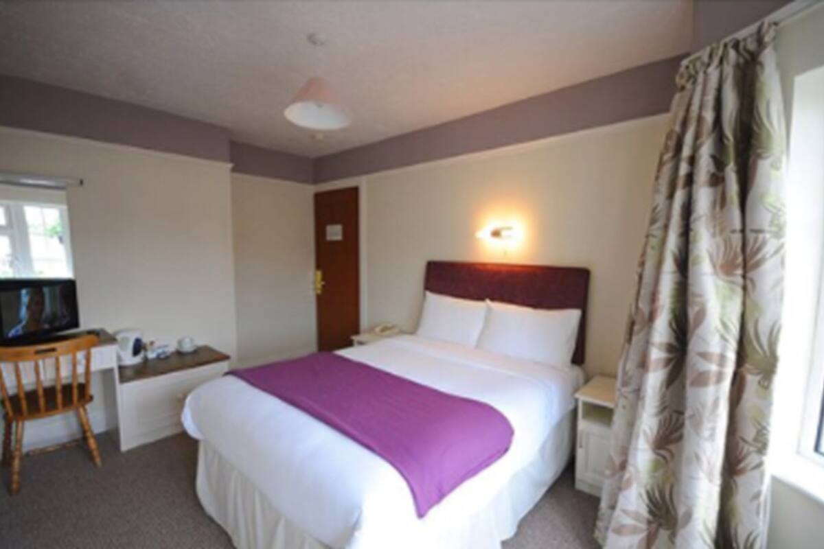 A hotel room with a bed, a desk with a chair, a television, a nightstand, and a window with curtains.