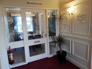 Un salón de desayunos con una puerta que da a un comedor, una planta en maceta y un extintor.