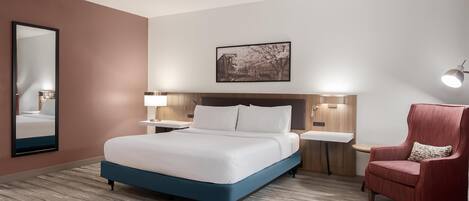 Une chambre d’hôtel avec un grand lit, un fauteuil rouge, une table de chevet avec une lampe, un miroir fixé au mur et un tableau encadré au-dessus du lit.