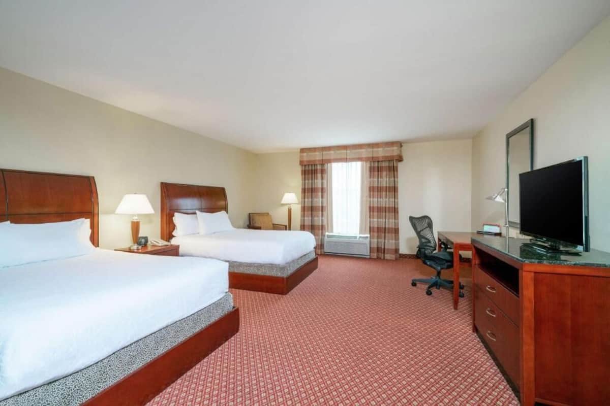 A hotel room with two beds, a desk, a TV, and a window with curtains.