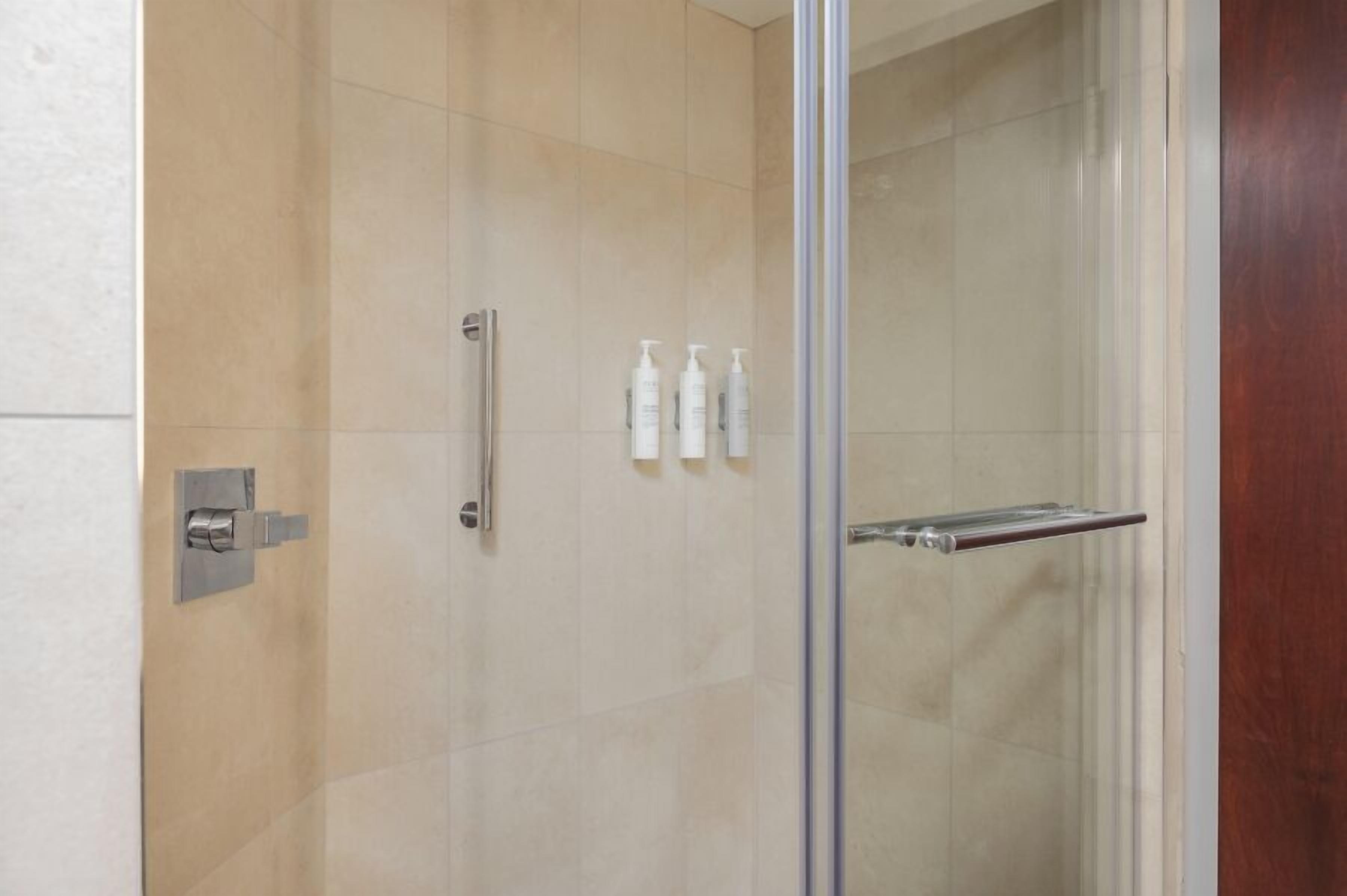 A glass-enclosed shower with a metallic handle, a towel rack, and three bottles of toiletries on the wall.