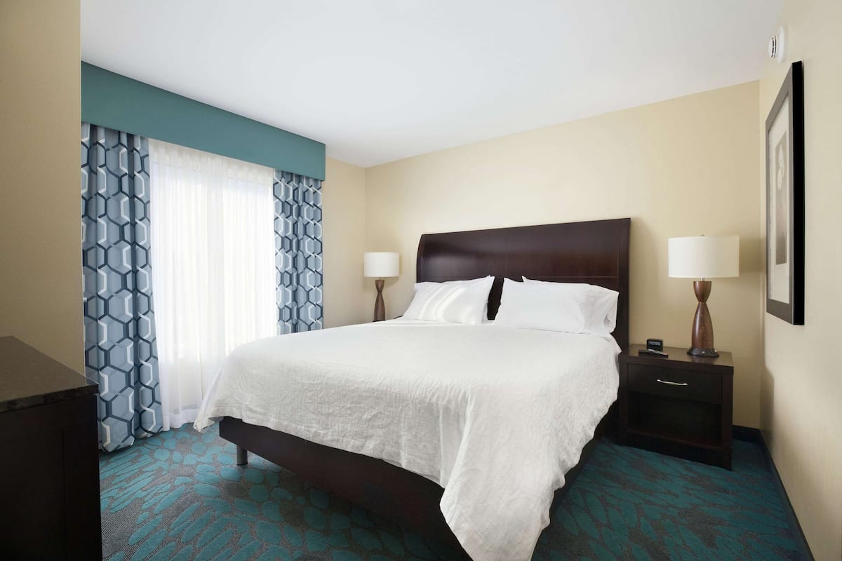 A hotel room with a large bed, two bedside tables with lamps, a dresser, and patterned curtains.