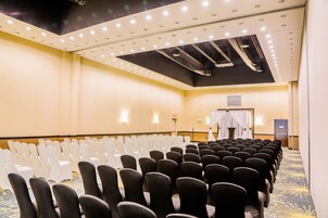 Un salón de conferencias con filas de sillas, un escenario con fondo blanco e iluminación empotrada en el techo.