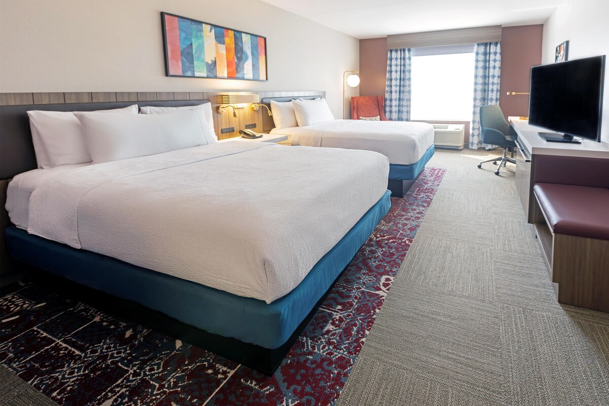 A hotel room with two beds, a desk with a chair, a television, and a large window with curtains.