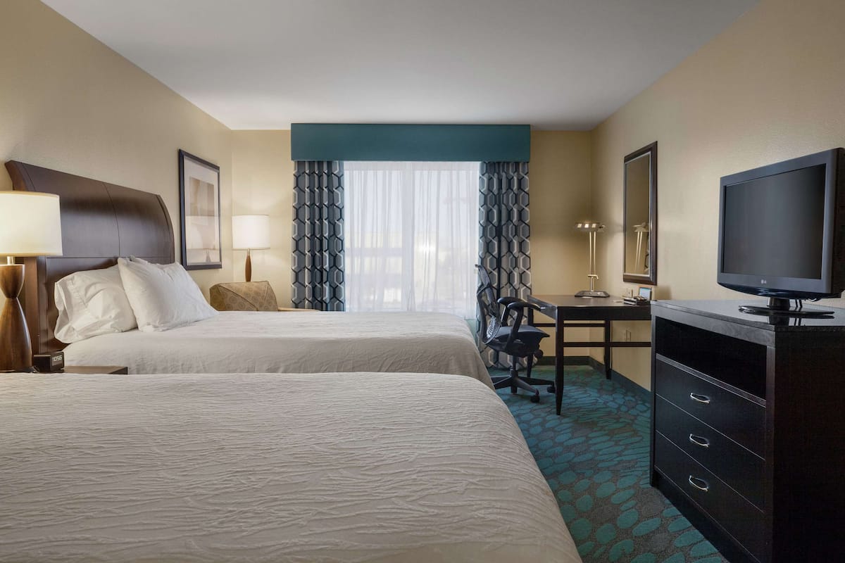 A hotel room with two beds, a desk with a chair, a television, and a large window with patterned curtains.