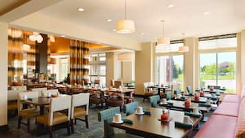 Ein Hotelrestaurant mit mehreren Tischen und Stühlen, großen Fenstern und Deckenleuchten.
