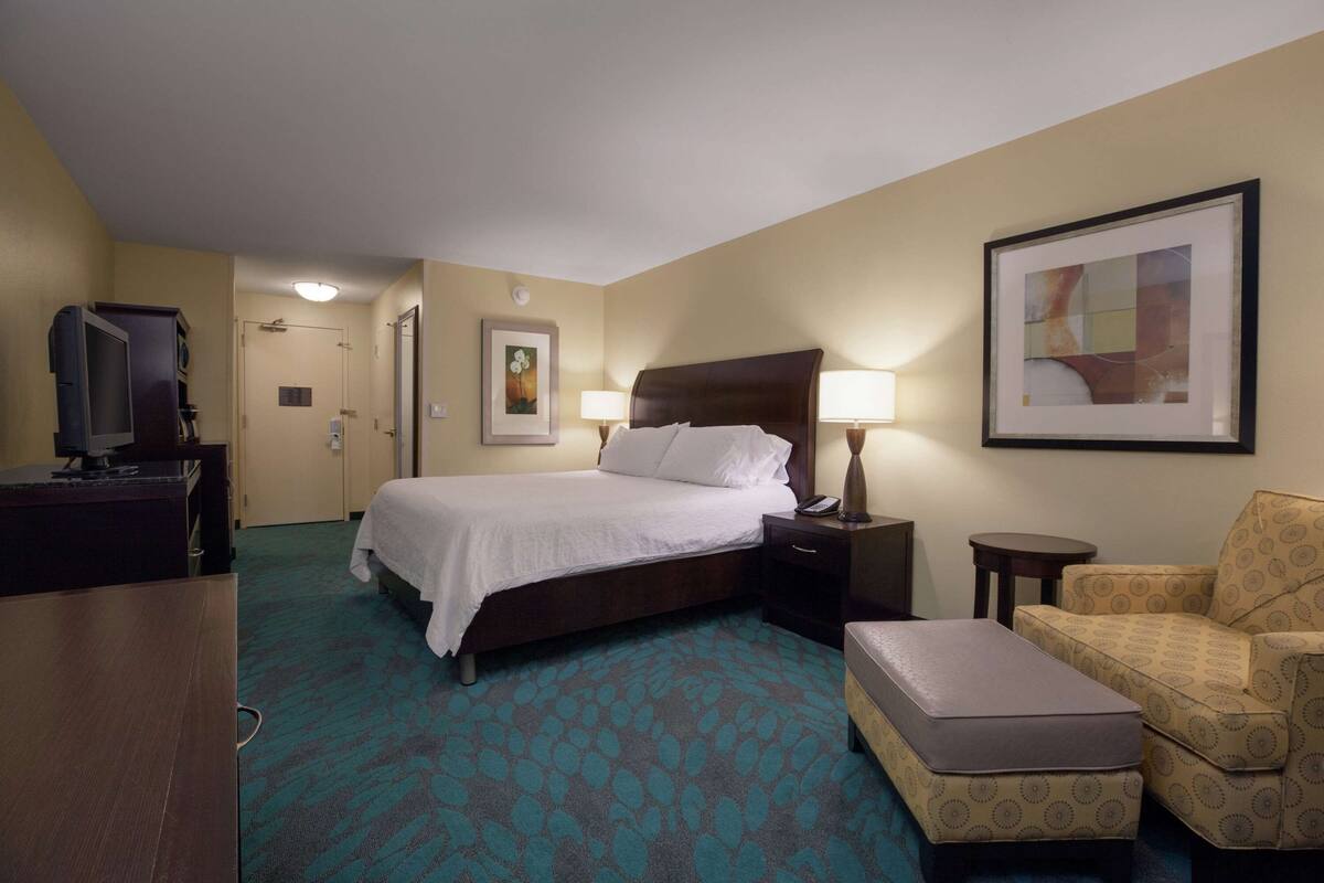 A hotel room with a bed, a desk with a television, a chair, a small table, and a lamp.