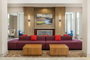 A modern living room with a large sectional sofa, two wooden side tables, a fireplace, and a large abstract painting above the fireplace. - Hilton Garden Inn Fargo (Fargo)