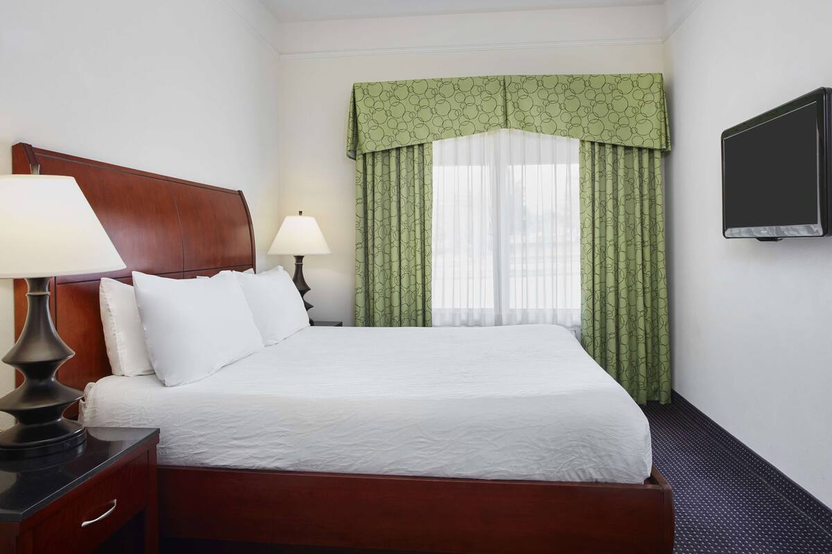 Une chambre d’hôtel avec une tête de lit en bois, un lit recouvert de linge de maison blanc, deux lampes de chevet, un téléviseur fixé au mur et des rideaux à motifs verts.