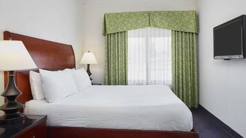 Une chambre d’hôtel avec une tête de lit en bois, un lit recouvert de linge de maison blanc, deux lampes de chevet, un téléviseur fixé au mur et des rideaux à motifs verts.
