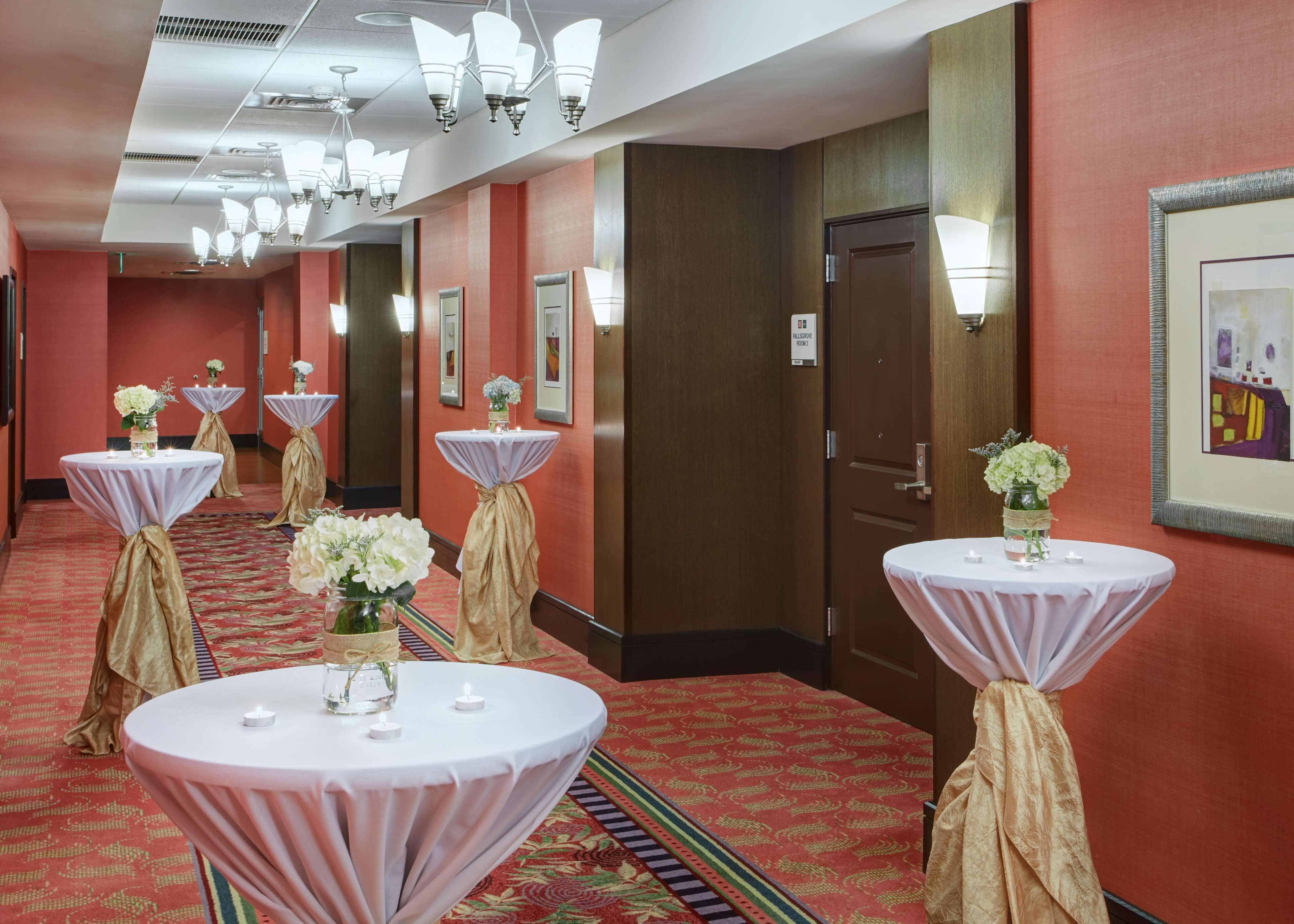A hotel corridor with round tables, each with a white tablecloth and a gold drape, along a red carpeted floor.