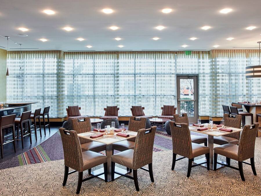 Uma sala de conferências de hotel com uma mesa grande e cadeiras, área de bar e janelas amplas com cortinas.