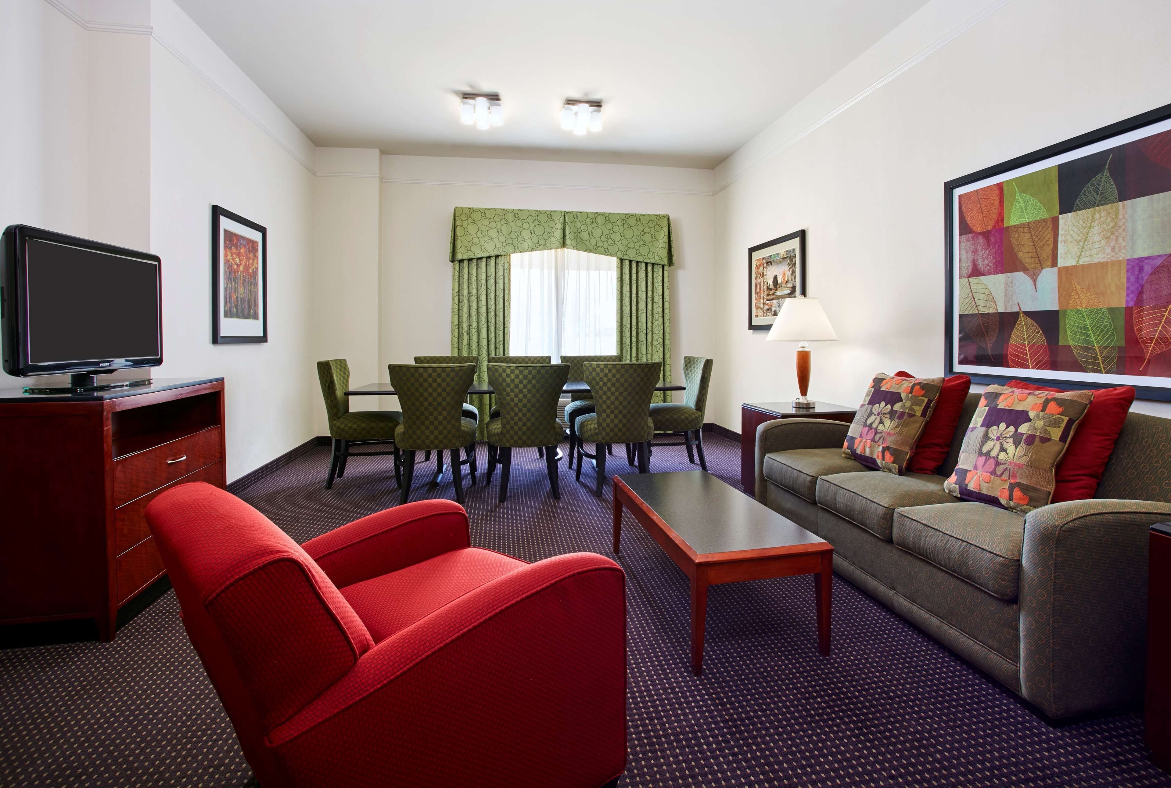 Una habitación de hotel con televisor, dos sillas rojas y verdes, un sofá, una mesa y un ventanal con cortinas.