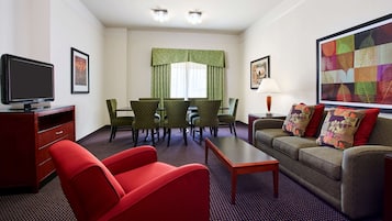 Una habitación de hotel con televisor, dos sillas rojas y verdes, un sofá, una mesa y un ventanal con cortinas.
