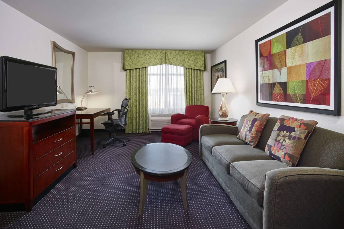 Une chambre d’hôtel équipée d’un bureau, d’une télévision, d’un canapé, d’un fauteuil rouge et d’une grande œuvre d’art encadrée accrochée au mur.