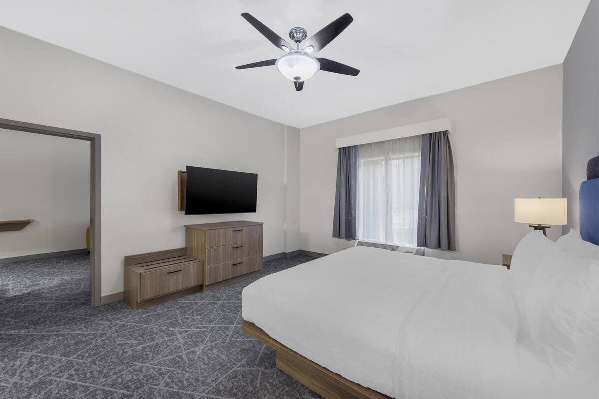 A hotel room with a bed, a flat-screen TV, a ceiling fan, a window with curtains, and a nightstand with a lamp.