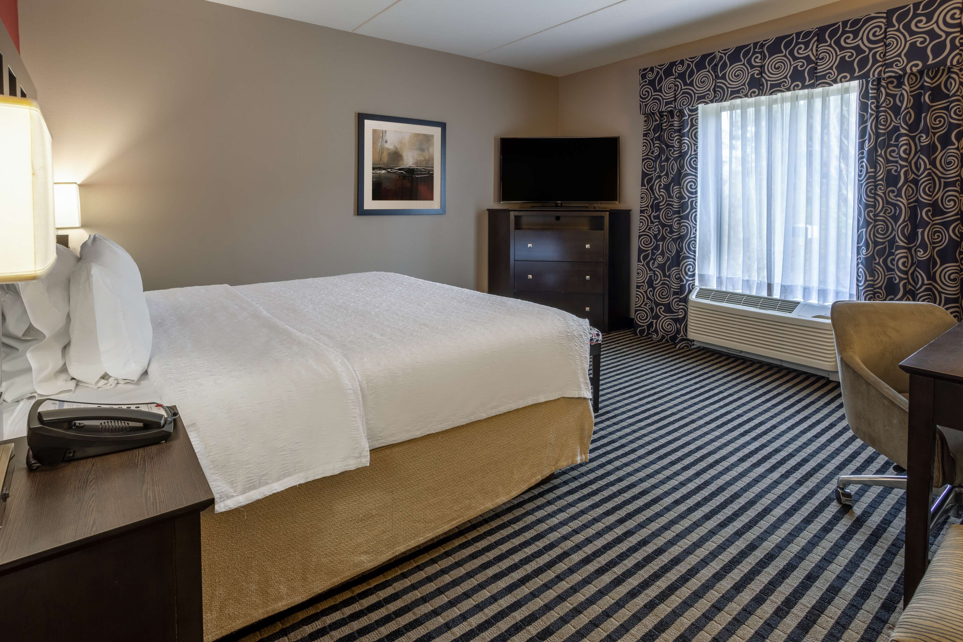 Una habitación de hotel con una cama, un escritorio con lámpara, televisor, una silla y una ventana con cortinas.