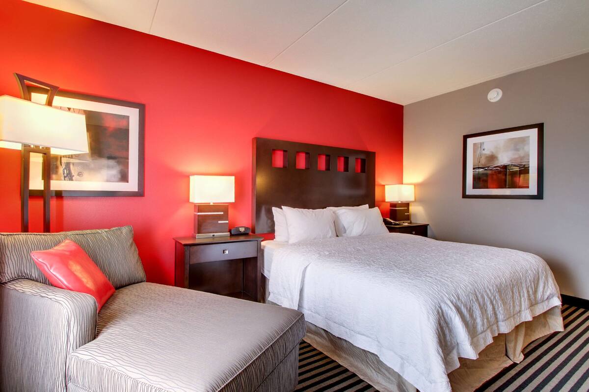 A hotel room with a red accent wall, a large bed with white bedding, a grey sofa with a red pillow, two bedside tables with lamps, and a framed picture on the wall.