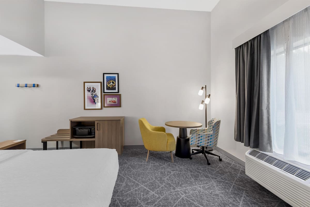 A hotel room with a bed, a desk, a microwave, a chair, a table, a lamp, and framed pictures on the wall.