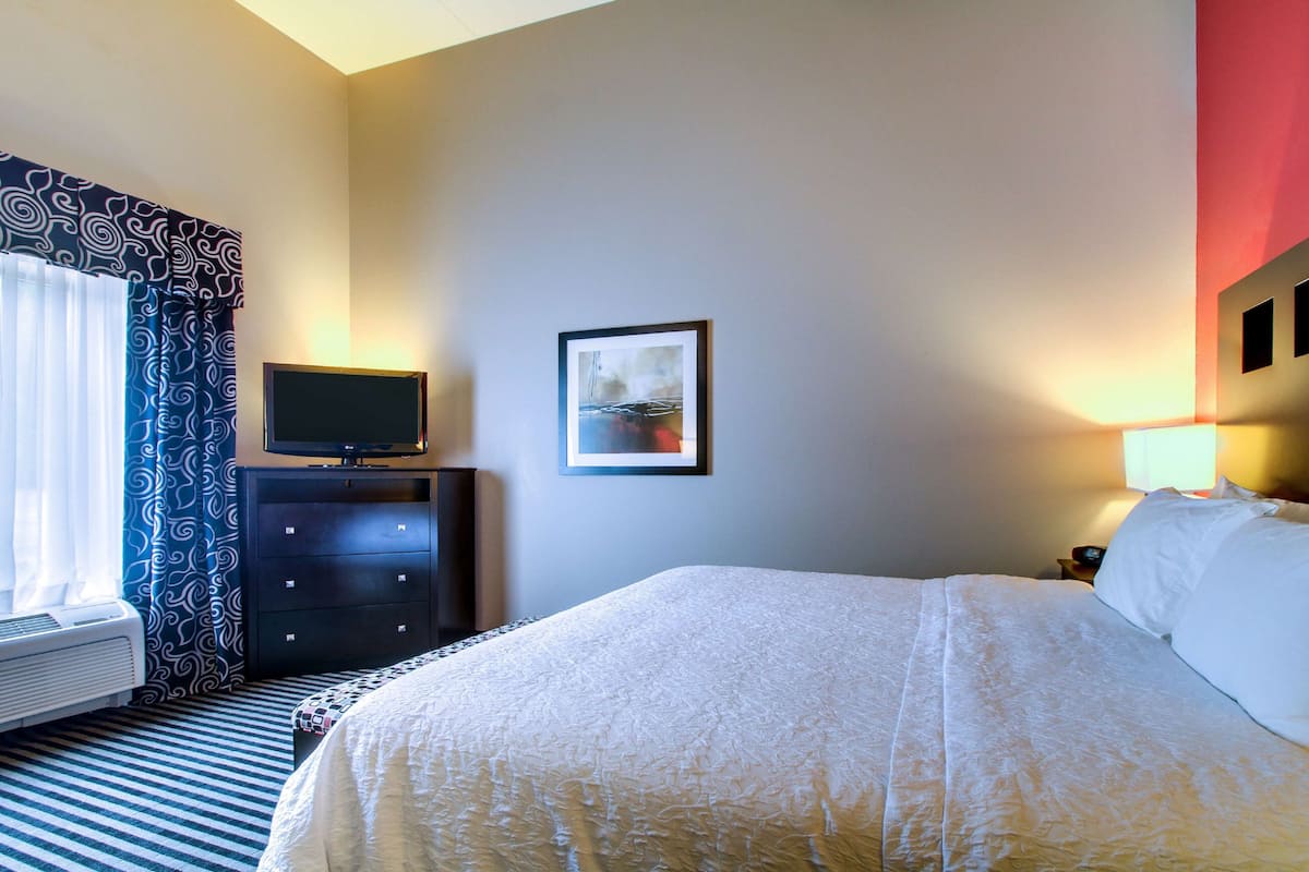 A hotel room with a bed, a TV on a dresser, a framed picture on the wall, and a window with curtains.