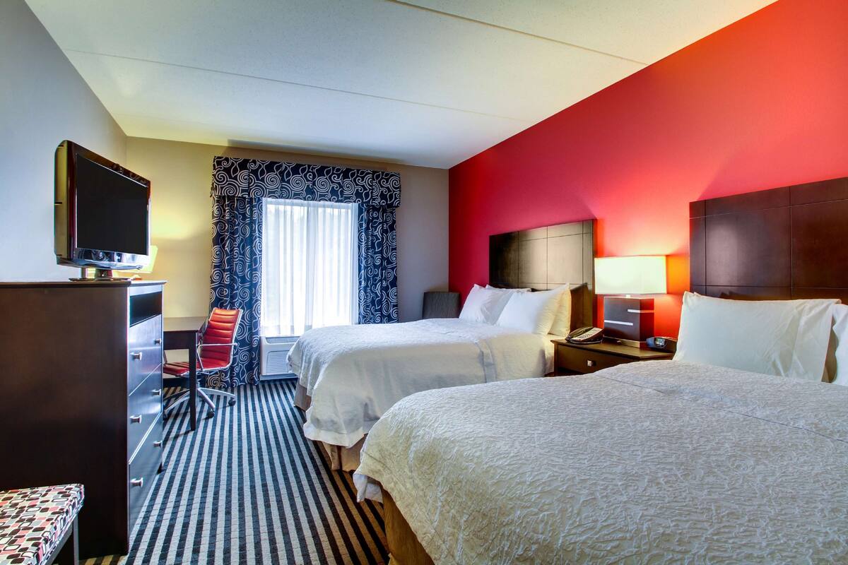 A hotel room with two beds, a desk, a TV, and a window with patterned curtains.