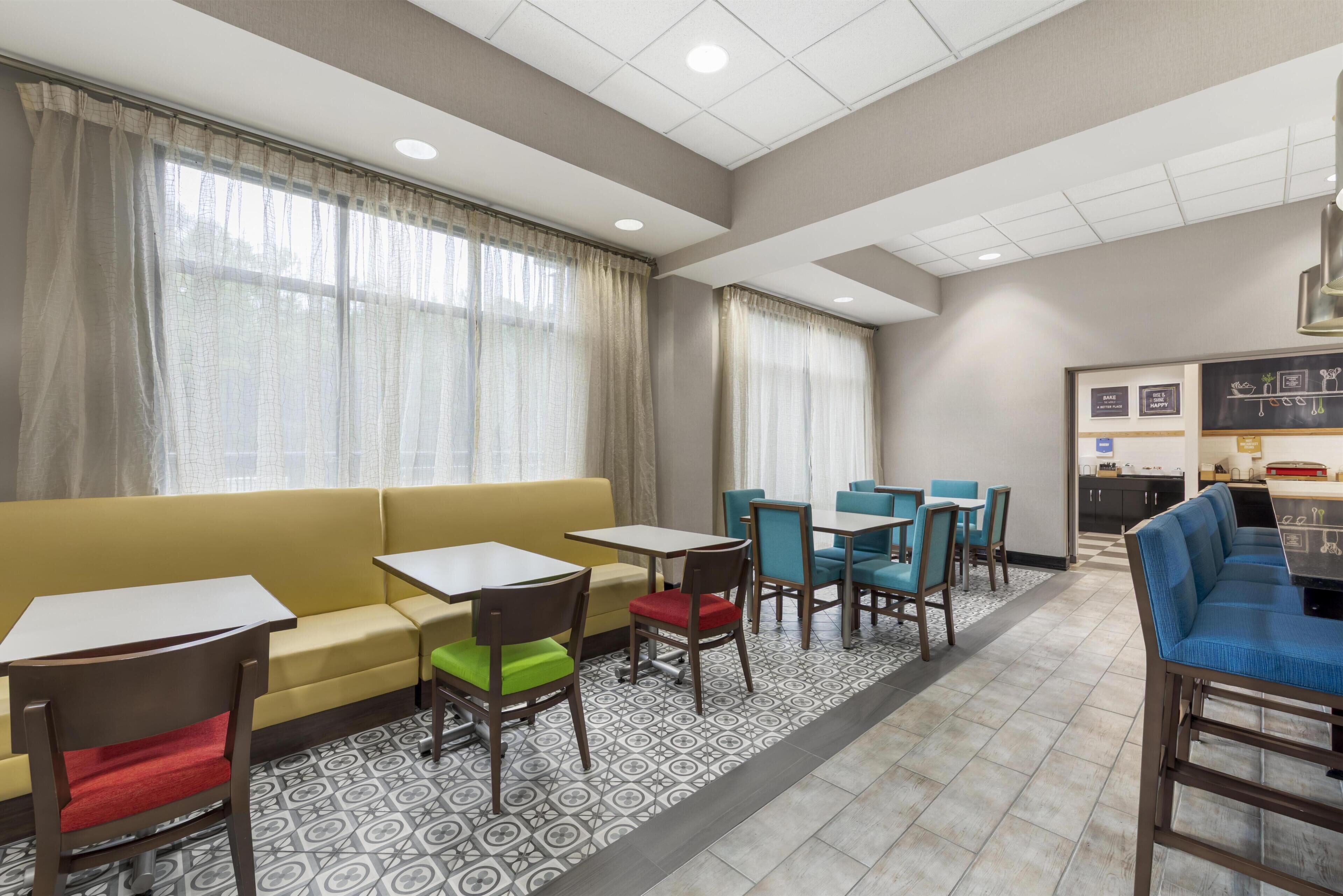 A hotel lobby with a yellow sofa, multiple tables and chairs, a kitchenette, and large windows with curtains.