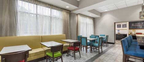 A hotel lobby with a yellow sofa, multiple tables and chairs, a kitchenette, and large windows with curtains.