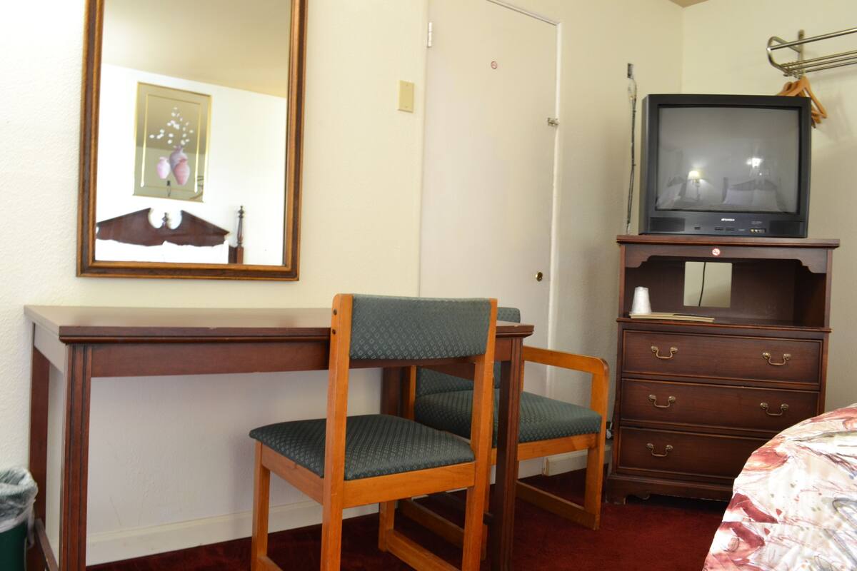 A hotel room with a wooden desk, a chair, a dresser, and a television on top of the dresser.