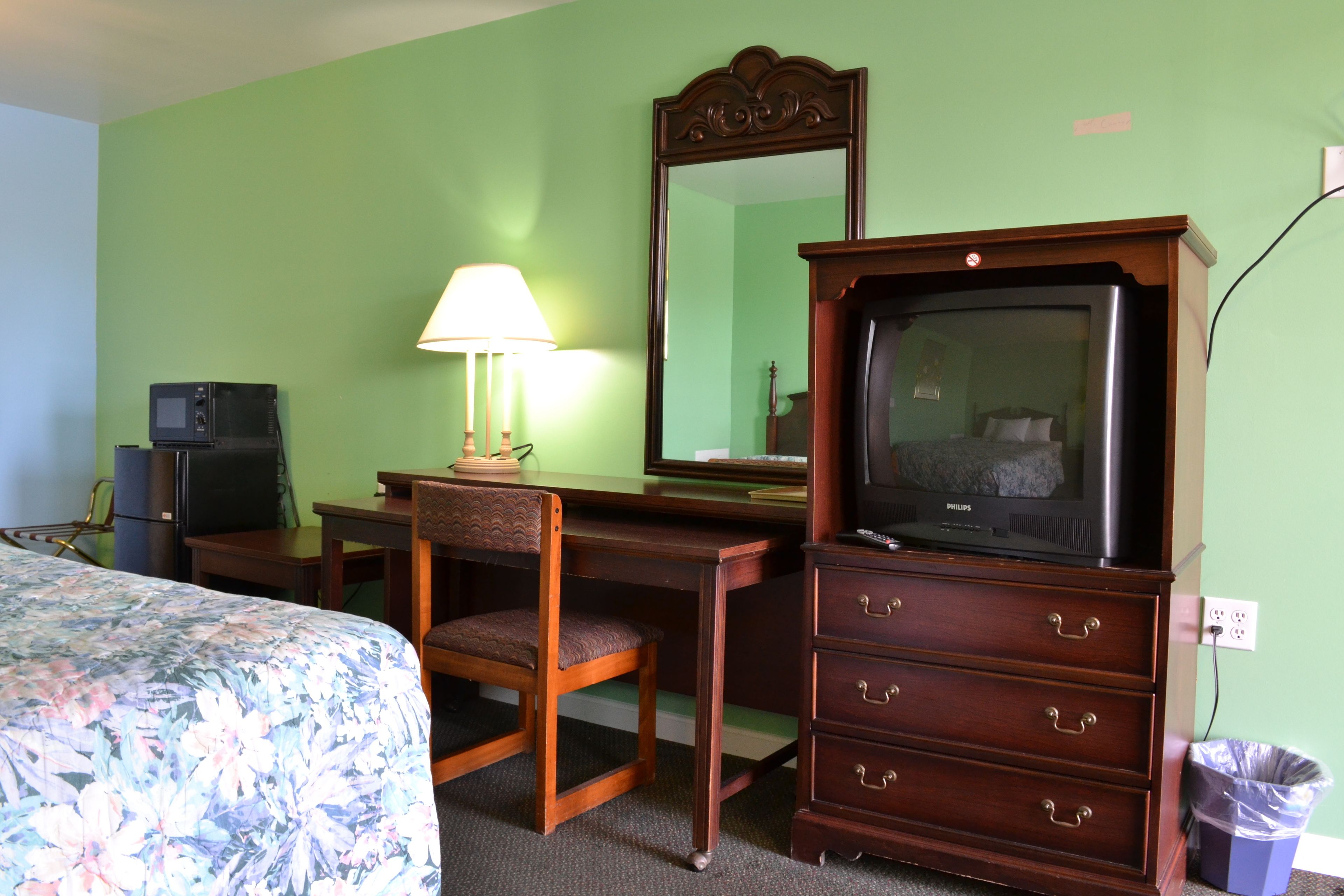 A hotel room with a bed, a desk with a lamp, a television, a dresser, and a trash can.