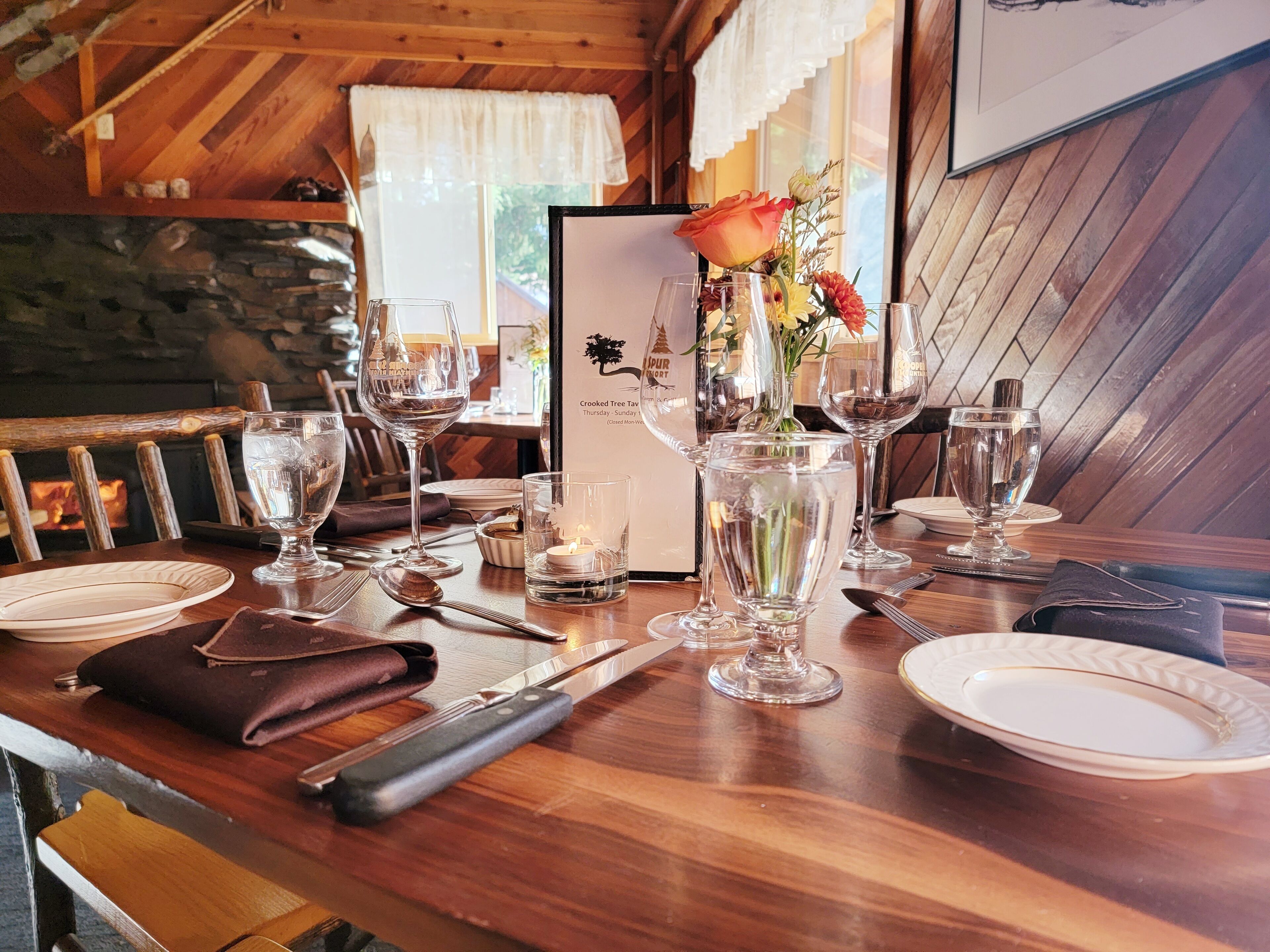 A dining table set with plates, cutlery, and wine glasses in a rustic wooden cabin with a fireplace and a framed menu.