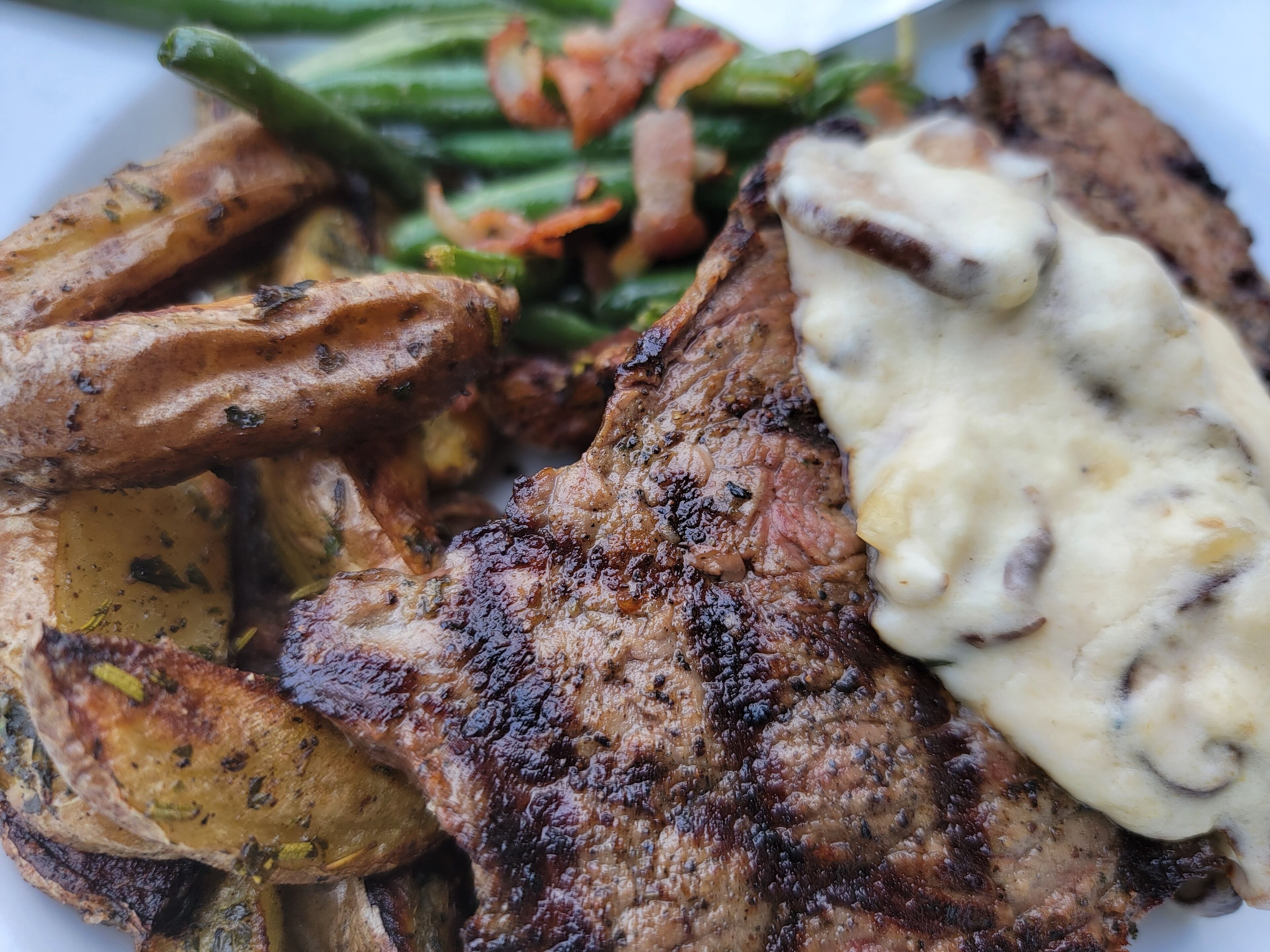 Un plato de comida con carne a la parrilla, papas asadas, judías verdes y salsa cremosa.
