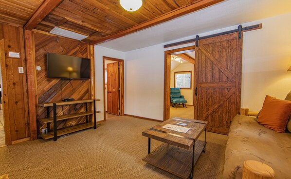 Ein gemütliches Wohnzimmer mit Holzbalkendecke, einem flachbildfernseher an einer Wand mit Fischgrätmuster, einer Schiebetür im Bauernhausstil, einem türkisfarbenen Sofa und einem Couchtisch mit Unterlagen.