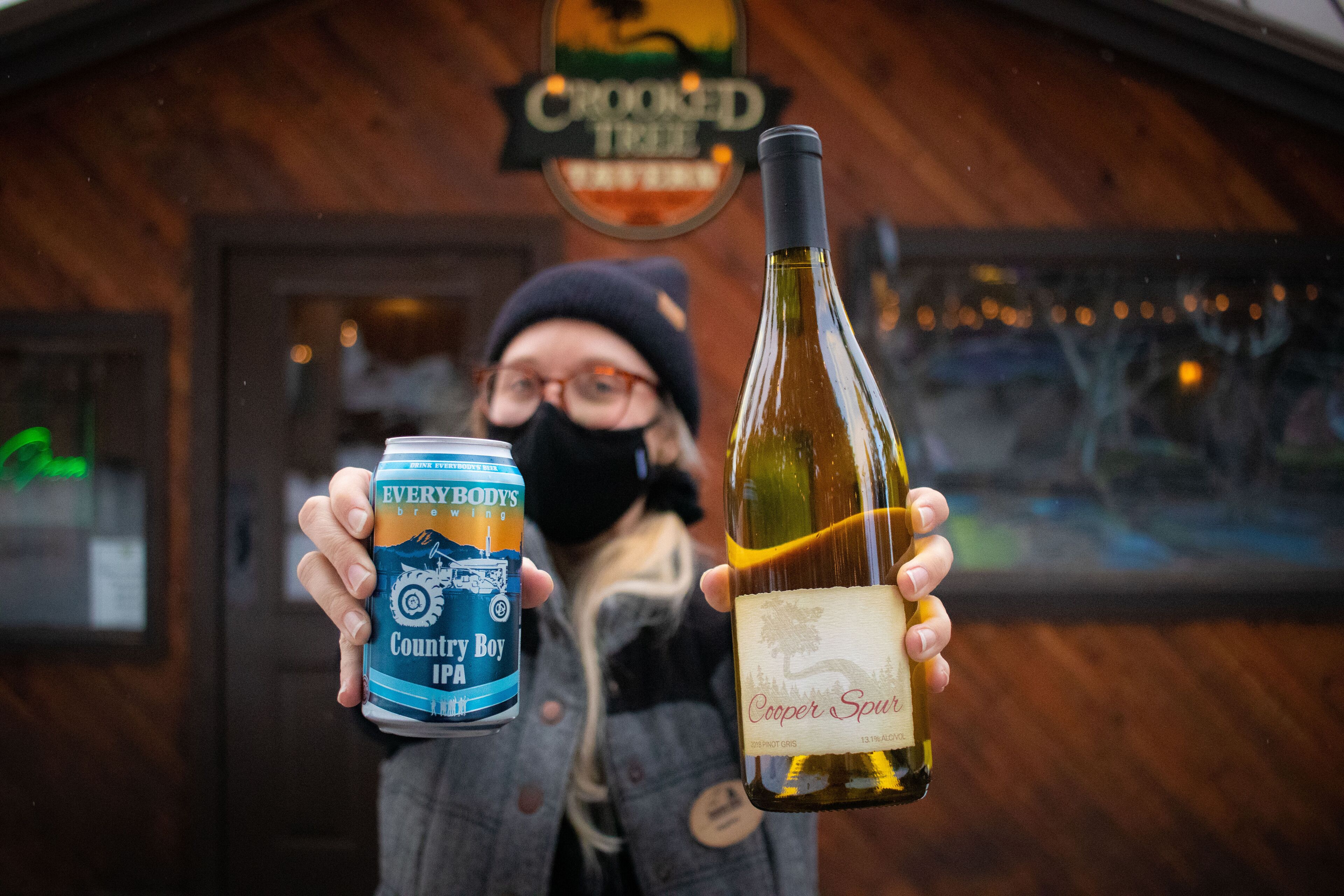 A person holding a can of beer and a bottle of wine in front of a building with a sign that reads "Crooked Tree".