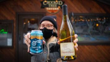 A person holding a can of beer and a bottle of wine in front of a building with a sign that reads "Crooked Tree".
